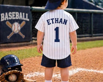 May include: A white baseball jersey with thin black pinstripes, the name ETHAN, and the number 1 in dark blue lettering on the back. The outfit includes dark blue shorts, white socks, white sneakers, and a dark blue baseball cap worn backward.