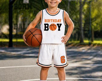 May include: A young boy wearing a white basketball uniform with orange and black trim, holding a basketball. The jersey says "BIRTHDAY BOY" with a basketball graphic. The shorts have the number "6". The boy is standing on a basketball court.