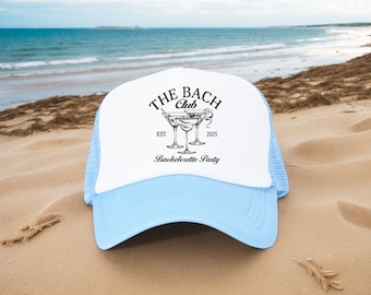 May include: A white and light blue trucker hat with the text "THE BACH Club" and "Bachelorette Party" in black. The hat features a graphic of three martini glasses and the year "2025". The hat is resting on a sandy beach with the ocean in the background.