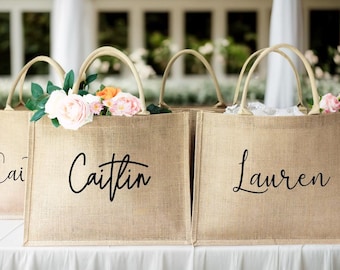 May include: Four burlap tote bags with cream-colored handles are displayed on a white surface. Each bag has a name in black script: "Cai", "Caitlin", and "Lauren". The bags are filled with flowers, including pink and orange roses.