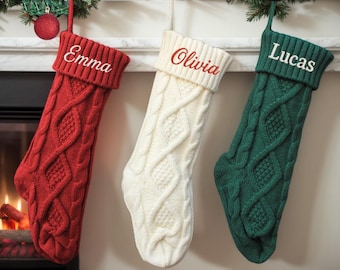 May include: Three cable-knit Christmas stockings in red, cream, and green hang from a mantel. Each stocking has a name embroidered on the cuff: "Emma" in white, "Olivia" in red, and "Lucas" in white. A red ornament hangs above the red stocking.