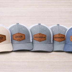 Op de afbeelding: Vijf baseballpetten met witte mesh-ruggen en gekleurde voorkanten. Elke pet heeft een bruine leren patch met tekst: "FATHER OF THE BRIDE", "GROOM", "GROOMSMAN", "GROOMSMAN" en "BEST MAN". De petten zijn beige, grijs en blauw.