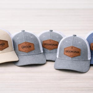 Peut inclure: Cinq casquettes de baseball avec des dos en filet et des écussons en cuir. Les casquettes sont de différentes couleurs : beige, gris et bleu. Les écussons en cuir portent les inscriptions "Father of the Bride", "Groom", "Groomsman" et "Best Man".