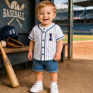 May include: A smiling toddler wearing a white baseball jersey with blue pinstripes and the number 1. The jersey is paired with denim shorts and white sneakers. A baseball bat, helmet, and glove are in the background, with the word "BASEBALL" visible.