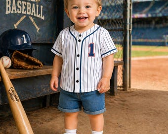 May include: A smiling toddler wearing a white baseball jersey with blue pinstripes and the number 1. The jersey is paired with denim shorts and white sneakers. A baseball bat, helmet, and glove are in the background, with the word "BASEBALL" visible.