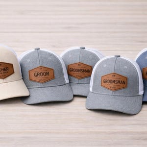 Op de afbeelding: Een verzameling baseballpetten met mesh-rug en leren patches. De petten zijn er in verschillende kleuren, waaronder beige, grijs en blauw. Elke patch heeft een andere titel, zoals "Vader van de bruid", "Bruidegom" en "Best Man."