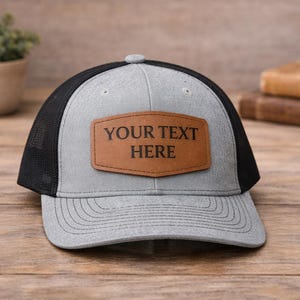 May include: A gray and black trucker hat with a leather patch that reads "YOUR TEXT HERE." The hat has a curved brim and a mesh back. The hat is sitting on a wooden surface.