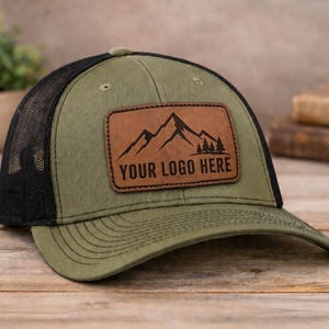 Peut inclure: Une casquette de baseball vert olive avec un dos en maille noire et un écusson rectangulaire en cuir marron sur le devant. L'écusson présente un graphique de montagnes noires, des sapins et le texte "YOUR LOGO HERE" gravé en gras.