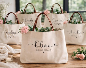 May include: Beige canvas tote bags with brown leather handles, each personalized with a name in elegant script and the word "Bridesmaid". The bags are filled with pink roses and greenery, perfect for a bridal party.