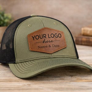 Puede incluir: Gorra de camionero verde oliva y negro con un parche de cuero marrón. El parche tiene el texto "YOUR LOGO here Name & Date". La gorra tiene una visera curva y una parte trasera de malla. Descansa sobre una superficie de madera.
