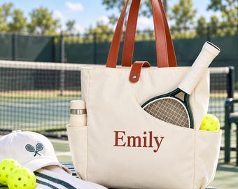 May include: A cream-colored canvas tote bag with brown leather handles and a snap closure. The bag features the name Emily printed in brown, a side pocket holding a water bottle, and a front pocket containing a pickleball paddle and ball.