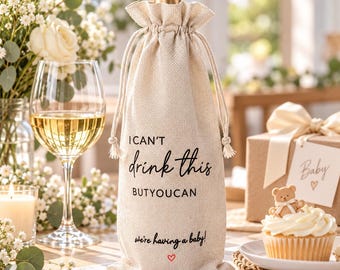 May include: A beige burlap wine bottle gift bag with a drawstring closure, featuring the black printed text "I CAN'T drink this BUTYOUCAN we're having a baby!" and a small red heart icon centered at the bottom of the fabric bag.