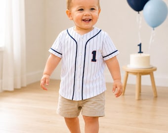 May include: A white short-sleeved baseball jersey with thin vertical navy blue pinstripes, a navy blue placket trim, and the number 1 in navy blue with a red outline on the chest, paired with plain tan shorts for a classic athletic look.