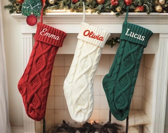 May include: Three cable-knit Christmas stockings in red, white, and green hang from a fireplace mantel. Each stocking is personalized with a name embroidered in a contrasting color: "Emma" in white, "Olivia" in red, and "Lucas" in white.