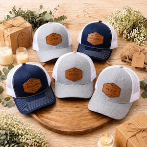 May include: Five trucker caps with personalised leather patches. The caps are in navy blue and grey with white mesh backs. The patches read "Groom" and "Groomsmen" with initials and dates. Wedding theme.