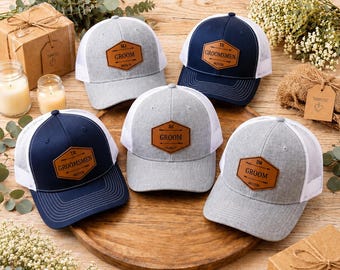May include: Five trucker hats with personalized leather patches. The hats are in navy blue and gray with white mesh backs. The patches read "Groom" and "Groomsmen" with initials and dates. Wedding theme.
