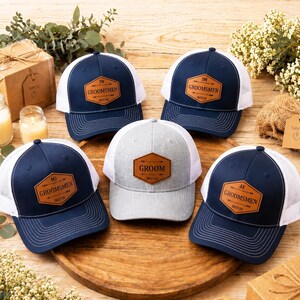 Pode incluir: Cinco bonés de beisebol com designs em malha azul marinho e branca são dispostos sobre uma superfície de madeira. Cada boné apresenta um patch de couro com texto, incluindo "GROOMSMEN" e iniciais. O boné central é cinza com "GROOM".