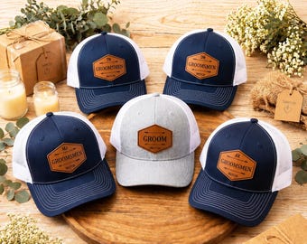 May include: Five baseball caps with navy blue and white mesh designs are arranged on a wooden surface. Each cap features a leather patch with text, including "GROOMSMEN" and initials. The central cap is gray with "GROOM" on its patch.
