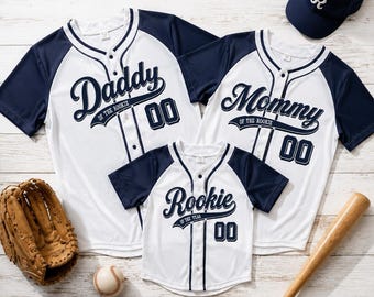 May include: Three white and navy blue baseball jerseys with button-fronts, featuring the text Daddy of the Rookie, Mommy of the Rookie, and Rookie of the Year with the number 00. Includes a navy blue baseball cap with an R, a glove, and a bat.