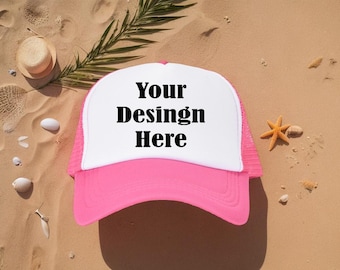 May include: A pink and white trucker hat with the text "Your Design Here" on the front. The hat is on a sandy surface with a straw hat, seashells, and a starfish.