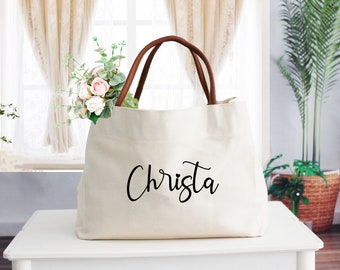 May include: A white canvas tote bag with brown leather handles. The bag has the name "Christa" printed on the front in black cursive.