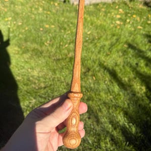 May include: A hand-turned wooden wand featuring a light brown grain pattern, a rounded pommel base, a textured grip section, and a long, slender, tapered shaft that extends to a pointed tip, showcasing natural wood textures and light brown tones.