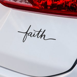 Op de afbeelding: Een witte auto met een zwarte vinyl sticker waarop in cursief schrift "faith" staat.