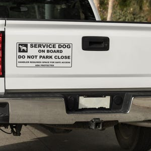May include: A white pickup truck's tailgate with a "SERVICE DOG ON BOARD" sticker. The sticker has a dog silhouette and text that reads "DO NOT PARK CLOSE" and "HANDLER REQUIRES SPACE FOR SAFE ACCESS ADA PROTECTED". The truck has a chrome bumper and red taillights.