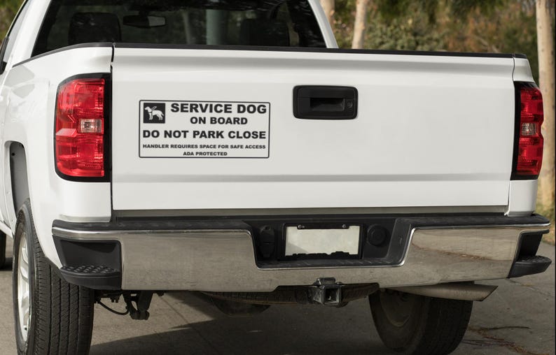 May include: A white pickup truck's tailgate with a "SERVICE DOG ON BOARD" sticker. The sticker has a dog silhouette and text that reads "DO NOT PARK CLOSE" and "HANDLER REQUIRES SPACE FOR SAFE ACCESS ADA PROTECTED". The truck has a chrome bumper and red taillights.