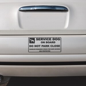 May include: A white car's rear with a rectangular sign that reads "SERVICE DOG ON BOARD" with a dog silhouette. The sign also states "DO NOT PARK CLOSE" and "HANDLER REQUIRES SPACE FOR SAFE ACCESS ADA PROTECTED."