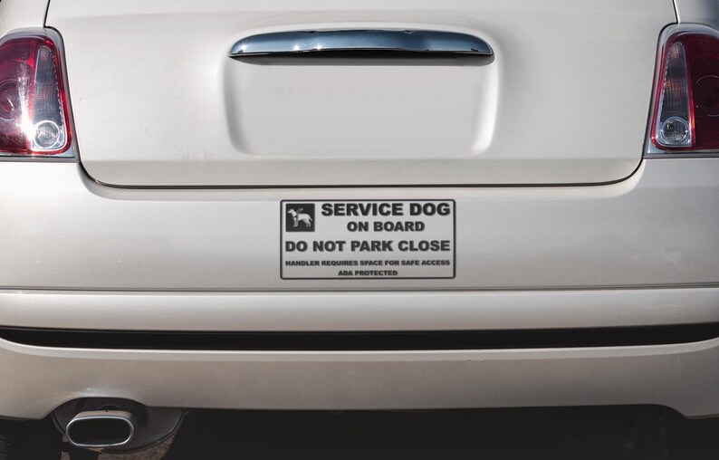 May include: A white car's rear with a rectangular sign that reads "SERVICE DOG ON BOARD" with a dog silhouette. The sign also states "DO NOT PARK CLOSE" and "HANDLER REQUIRES SPACE FOR SAFE ACCESS ADA PROTECTED."