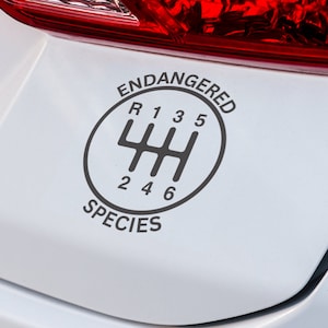 Peut inclure: Un autocollant noir et gris représentant un schéma de changement de vitesse de transmission manuelle. L'autocollant indique "ENDANGERED SPECIES" dans un design circulaire autour du schéma de changement de vitesse, avec les chiffres 1, 2, 3, 4, 5 et 6 visibles.