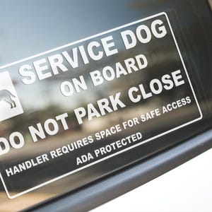 May include: A rectangular white sticker on a car window reads "SERVICE DOG ON BOARD DO NOT PARK CLOSE." The sticker includes a graphic of a dog and states "HANDLER REQUIRES SPACE FOR SAFE ACCESS ADA PROTECTED."