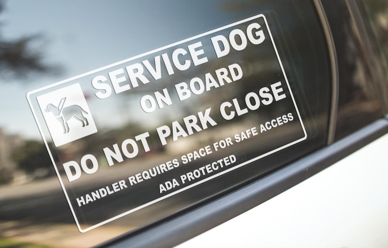 May include: A rectangular white sticker on a car window reads "SERVICE DOG ON BOARD DO NOT PARK CLOSE." The sticker includes a graphic of a dog and states "HANDLER REQUIRES SPACE FOR SAFE ACCESS ADA PROTECTED."