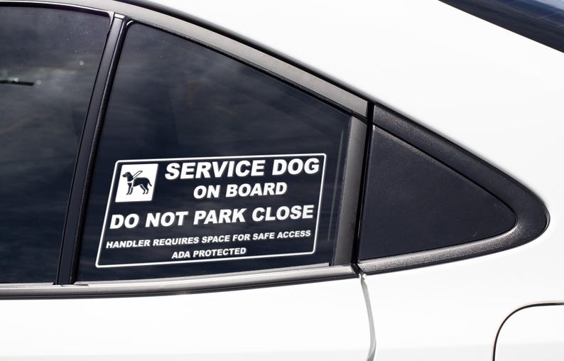 May include: A white rectangular sticker on a car window reads "SERVICE DOG ON BOARD DO NOT PARK CLOSE." The sticker includes a graphic of a dog and the text "HANDLER REQUIRES SPACE FOR SAFE ACCESS ADA PROTECTED."
