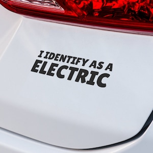 May include: A black and white bumper sticker with the text "I IDENTIFY AS A ELECTRIC" in a bold, sans-serif font. The sticker is affixed to the white rear of a vehicle, near the red taillight.