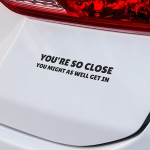 May include: A white car with a black vinyl sticker that reads "You're so close you might as well get in."