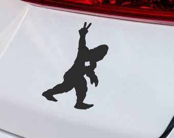 May include: Black silhouette of a person making a peace sign with their hand, with a bullet hole in their chest. The silhouette is on a white car.