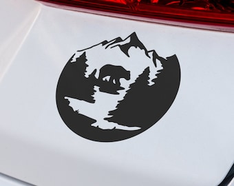 Könnte beinhalten: Schwarz-weißer Silhouettenaufkleber eines Bären, der durch einen Wald mit Bergen im Hintergrund geht. Der Aufkleber befindet sich auf einem weißen Auto.