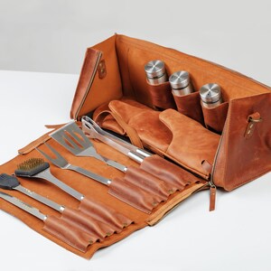 Op de afbeelding: Een bruine leren draagtas met een ritssluiting, die een set grillgereedschap bevat, waaronder een spatel, tang, grillborstel en een mes. De tas heeft ook vakken voor drie kleine flessen.