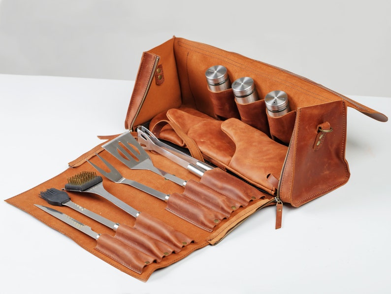 Op de afbeelding: Een bruine leren draagtas met een ritssluiting, die een set grillgereedschap bevat, waaronder een spatel, tang, grillborstel en een mes. De tas heeft ook vakken voor drie kleine flessen.