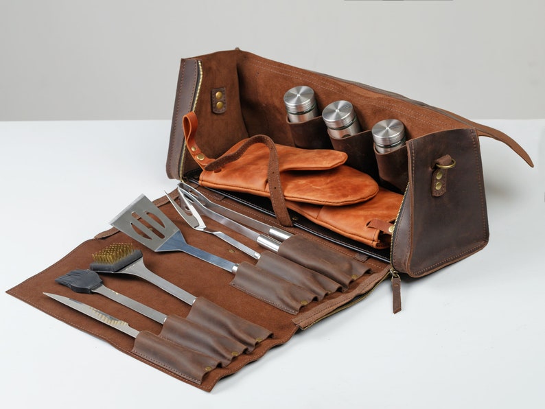 Op de afbeelding: Een bruine leren grillset met een draagtas met ritssluiting. De set bevat een spatel, tang, grillborstel, vork en mes. De tas heeft ook vakken voor drie kleine flessen en een paar leren grillhandschoenen.
