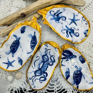 Könnte beinhalten: Vier weiße Keramik-Austernmuschel-Schalen mit goldenen Rändern. Jede Schale zeigt ein anderes blaues und weißes nautisches Design: einen Oktopus, einen Seestern und zwei Meeresschildkröten.