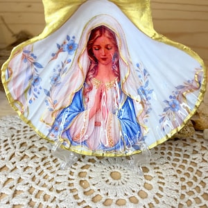 Puede incluir: Una concha de vieira decorativa con una ilustraci&oacute;n central de una mujer de pelo largo con velo blanco y t&uacute;nica azul, con las manos juntas en oraci&oacute;n. La concha est&aacute; pintada con bordes dorados y delicados detalles florales azules sobre fondo blanco.