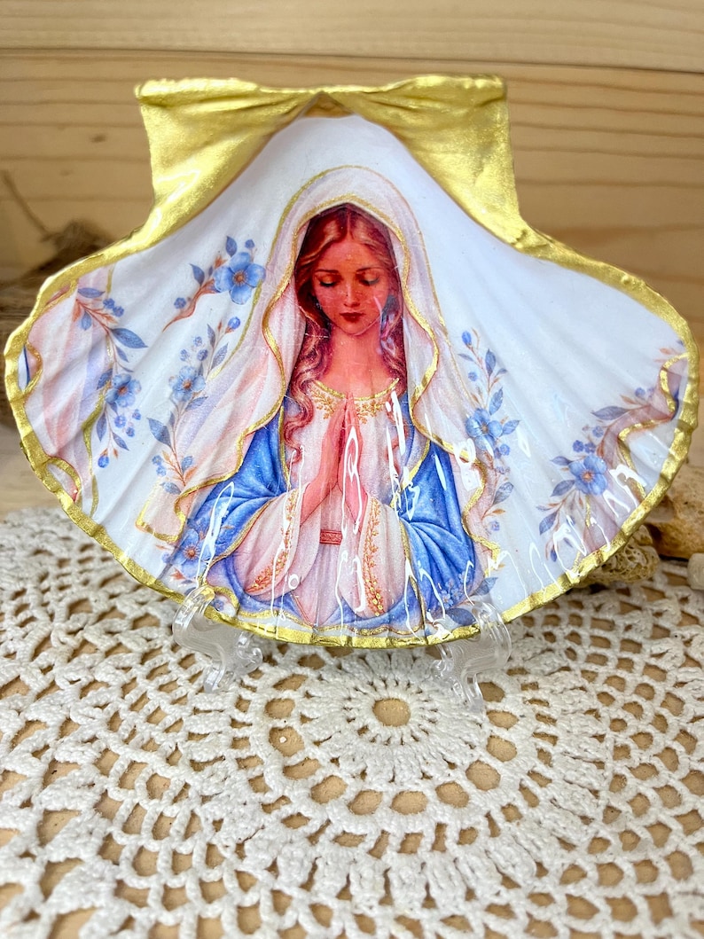 Puede incluir: Una concha de vieira decorativa con una ilustraci&oacute;n central de una mujer de pelo largo con velo blanco y t&uacute;nica azul, con las manos juntas en oraci&oacute;n. La concha est&aacute; pintada con bordes dorados y delicados detalles florales azules sobre fondo blanco.