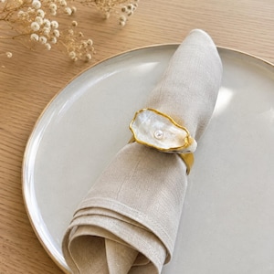 Può includere: Un tovagliolo di lino beige arrotolato e fissato con un portatovagliolo caratterizzato da un frammento di conchiglia di ostrica bianca iridescente con bordo dorato e una piccola perla bianca centrale, poggiato su un piatto in ceramica grigio chiaro.