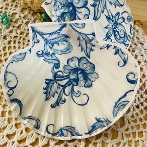 Peut inclure: Deux coquilles Saint-Jacques blanches avec des motifs floraux bleus. Les coquilles ont une finition brillante et sont décorées de motifs bleus complexes de fleurs et de feuilles. Les coquilles sont sur une surface crochetée blanche.