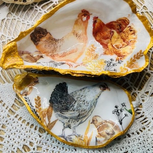 Puede incluir: Dos platos decorativos de concha de ostra con ribete dorado. Cada plato presenta un diseño diferente de acuarela de pollos y pollitos. Un plato tiene un diseño de pollo blanco, marrón y amarillo, mientras que el otro tiene un diseño de pollo negro, blanco y gris.