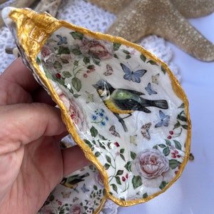Könnte beinhalten: Eine weiße Austernschalen mit goldenem Rand, verziert mit einem Blumenmuster, Schmetterlingen und einem kleinen Vogel. Die Schale ist eine dekorative Schale oder ein Schmuckhalter.