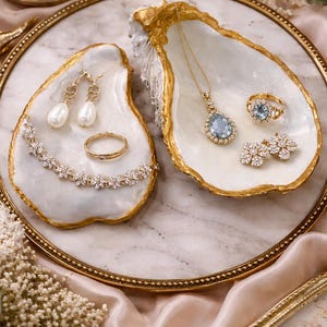 Könnte beinhalten: Elegante Schmuckpräsentation auf einem goldgerahmten Tablett. Zwei muschelförmige Schalen präsentieren Schmuck. Eine enthält eine Perlenkette, Ohrringe und einen Ring. Die andere zeigt eine Halskette mit Anhänger, einen Ring und Ohrringe mit blauen Edelsteinen.